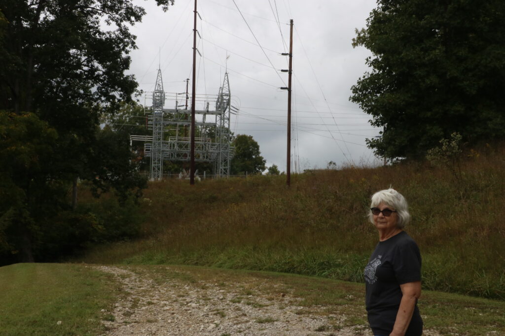 Brenda Campbell poses on her property, the background showing the large, metal electrical substation powering the new facility by Artemis Power Tech.