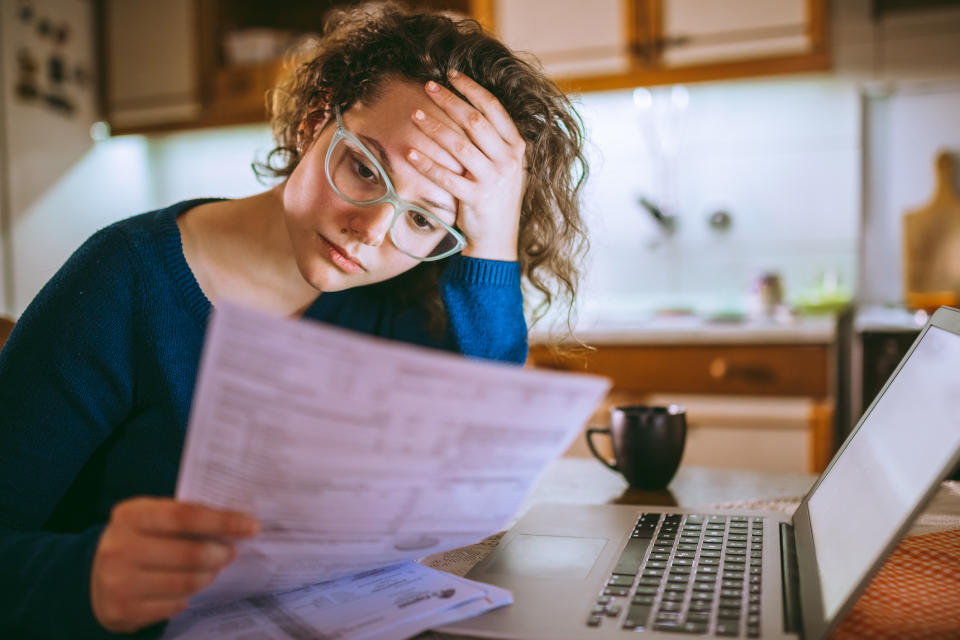 Person wearing glasses and sweater appears stressed while reviewing financial documents next to a laptop in a kitchen