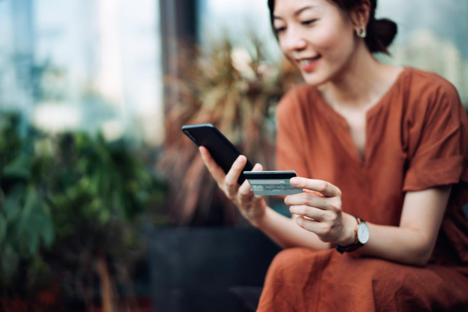 A woman in casual attire smiles while using a smartphone and holding a credit card, suggesting online shopping or mobile banking