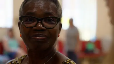 An older woman with gray hair named Vadney, wearing black-rimmed glasses and red earrings